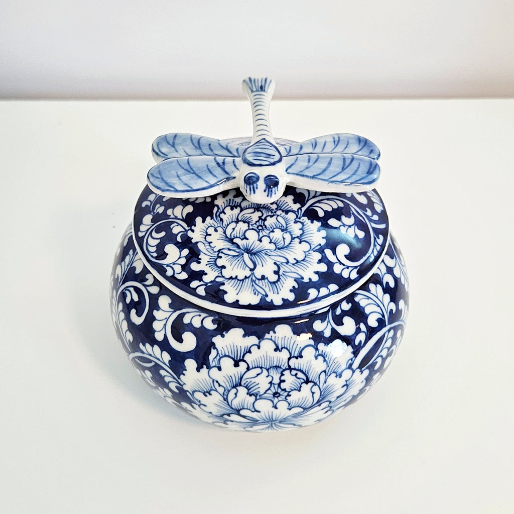 A round blue and white porcelain lidded jar decorated with floral patterns, topped with a dragonfly-shaped handle, set against a plain white background.