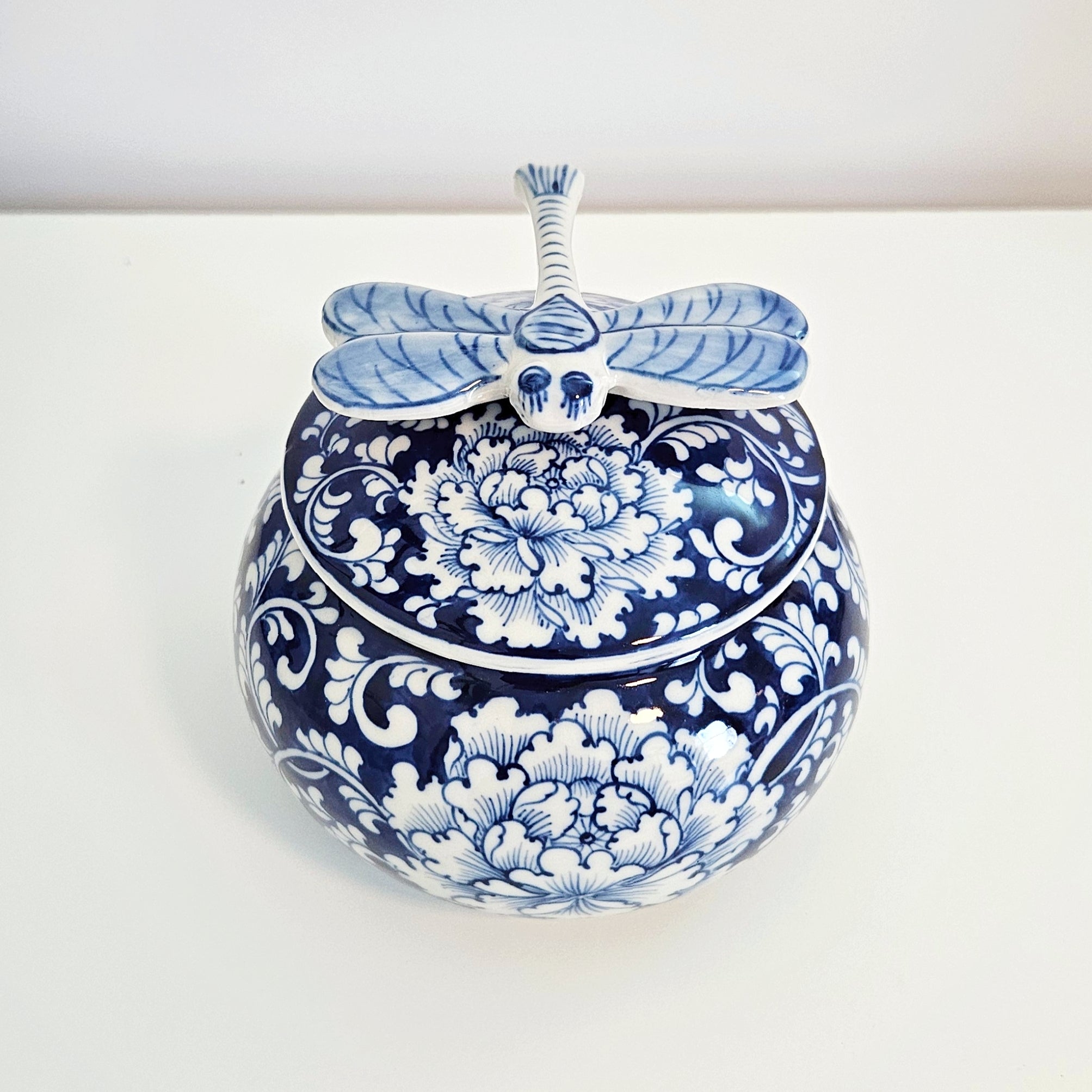 A round blue and white porcelain lidded jar decorated with floral patterns, topped with a dragonfly-shaped handle, set against a plain white background.
