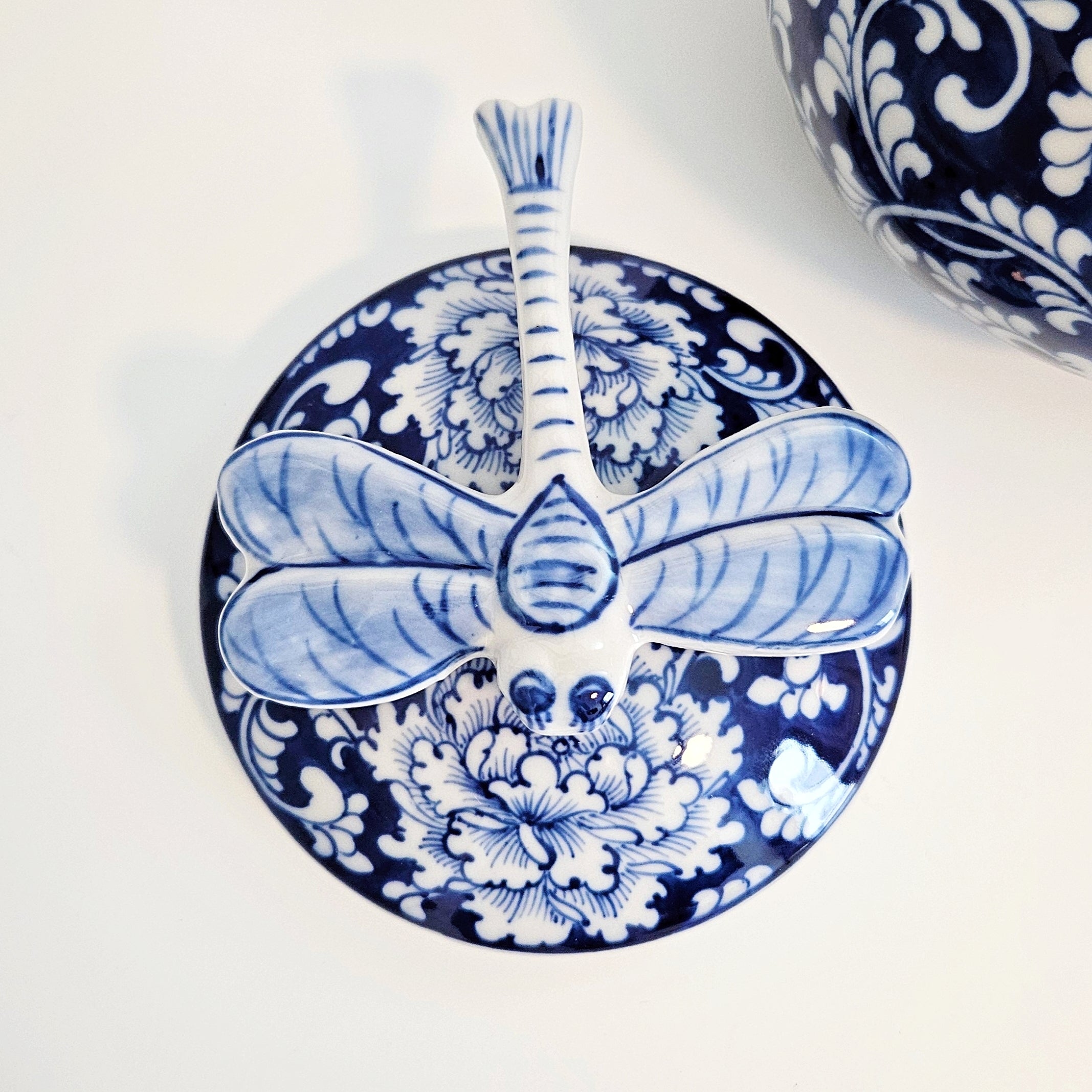 A blue and white ceramic dragonfly-shaped lid, photographed on a clean white background with part of another similar item visible in the upper right.