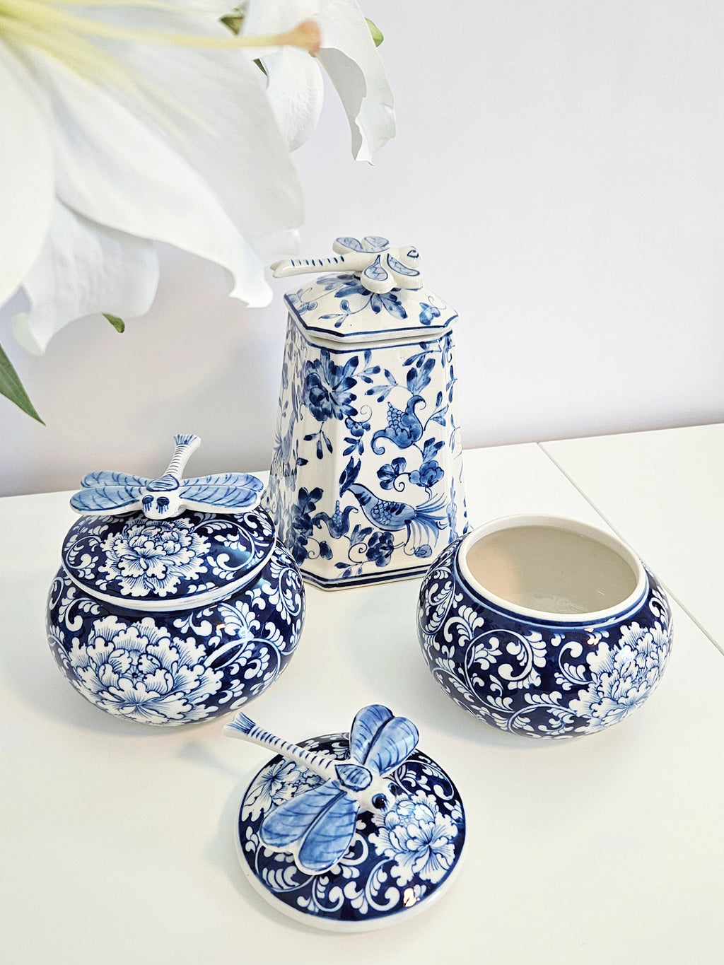 two blue and white chinoiserie porcelain jars with dragonfly lid placing next to a tall square porcelain jar with the same dragonfly design lid, all are putting on a white table
